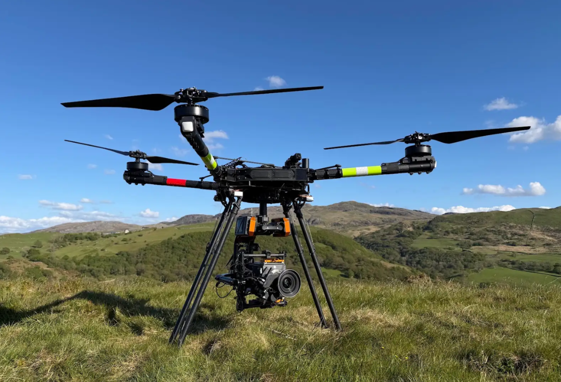Professional heavy-lift cinema drone equipped with a high-end camera rig standing on a grassy hill, used for cinematic drone filming against a scenic mountain backdrop.