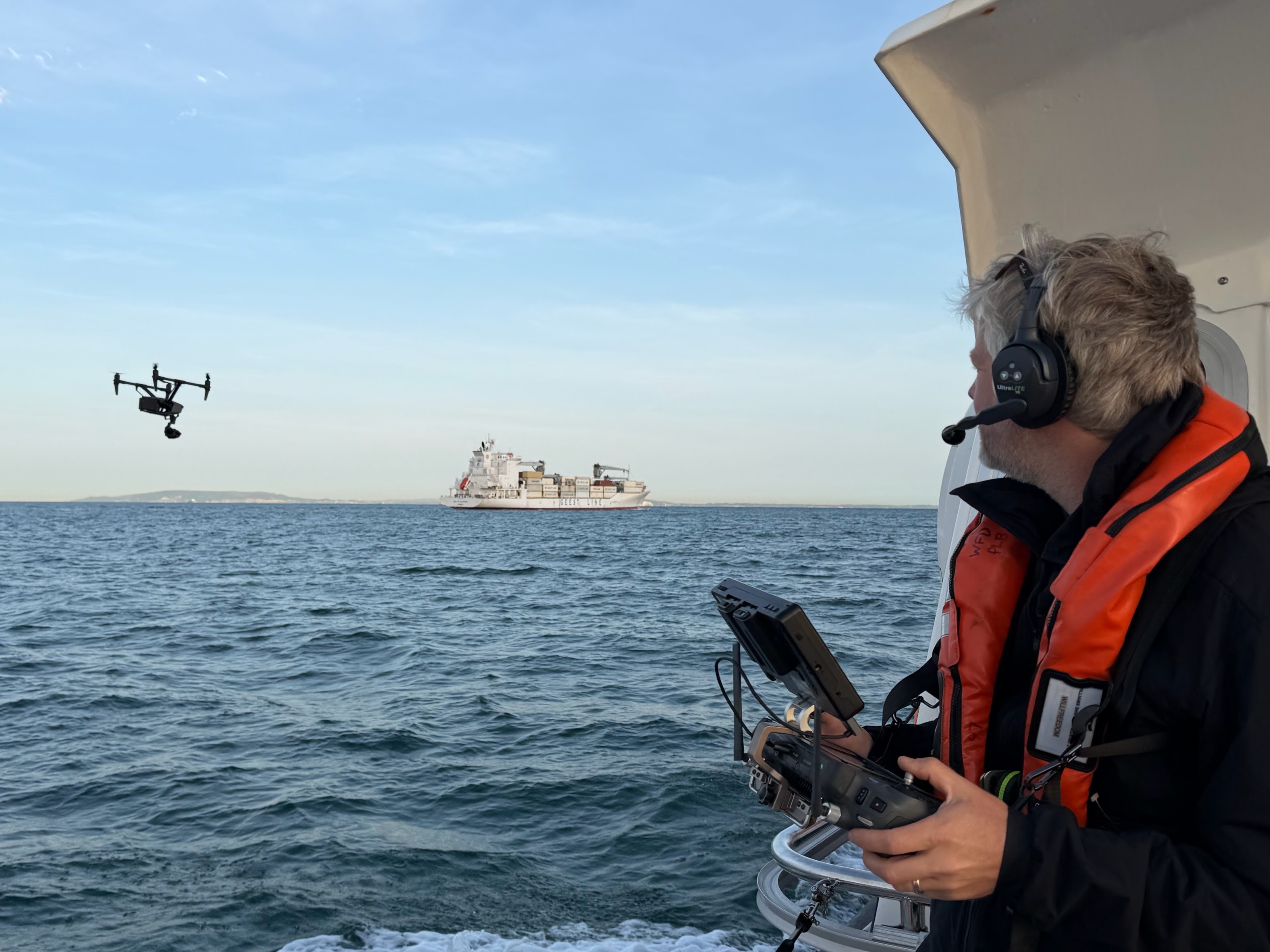 Drone videography team operating a camera-equipped drone over the sea near a cargo ship during a professional filming shoot.